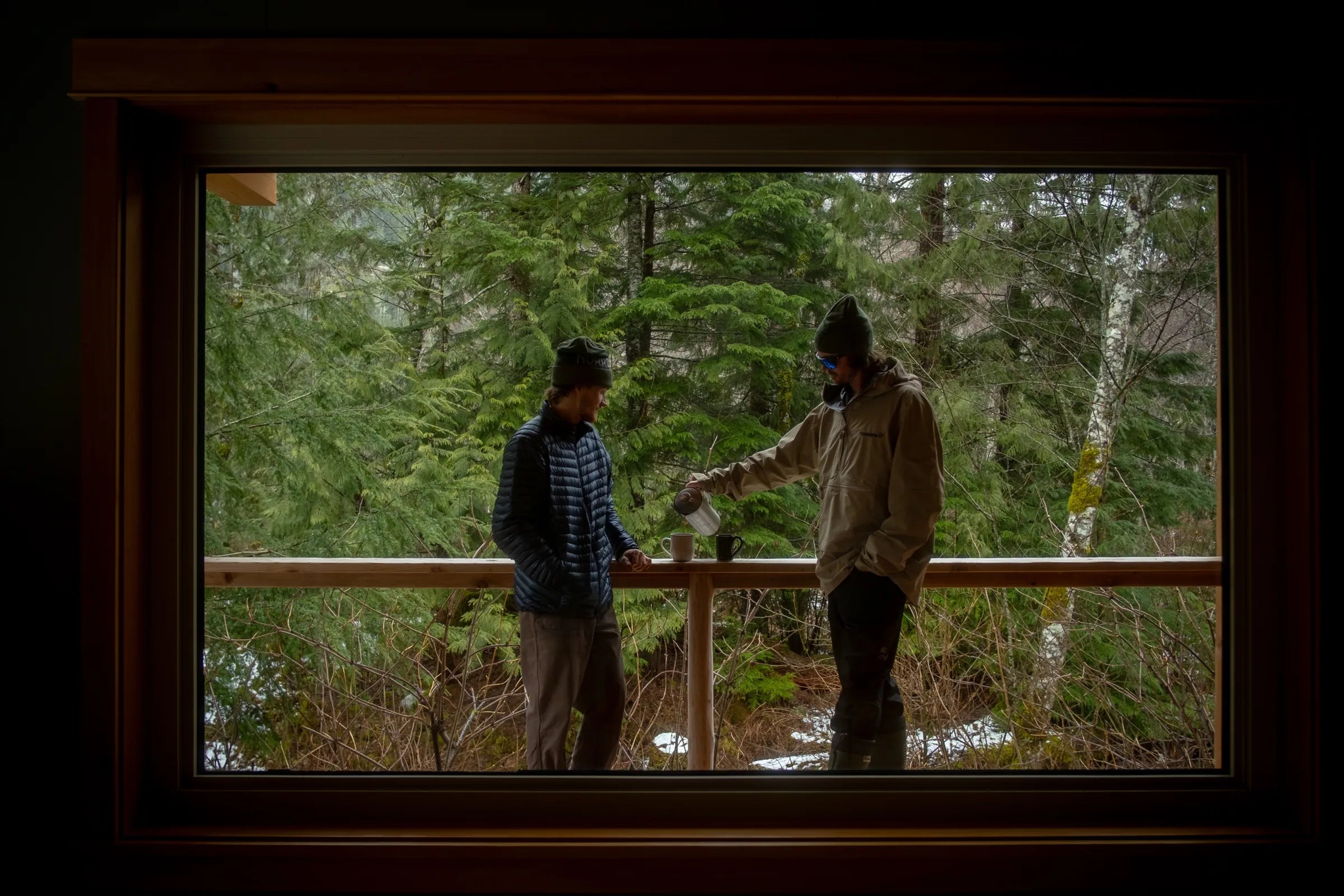 Two skiers on a lodge deck, one pouring coffee from a flask into a mug, forest behind